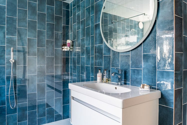 Modern bathroom design featuring vertical blue subway tiles, a white vanity unit, and a large round backlit mirror.